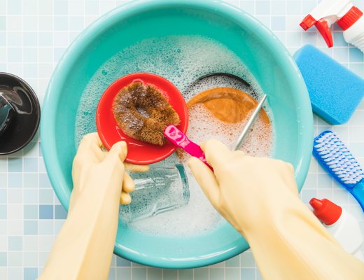 Two gloved hands cleaning various dishes with soapy water and a soft scrub brush.