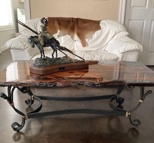 A live-edge wooden epoxy coffee table with elaborate metal legs that are linked together. A figure of a man riding a horse rests atop the table.