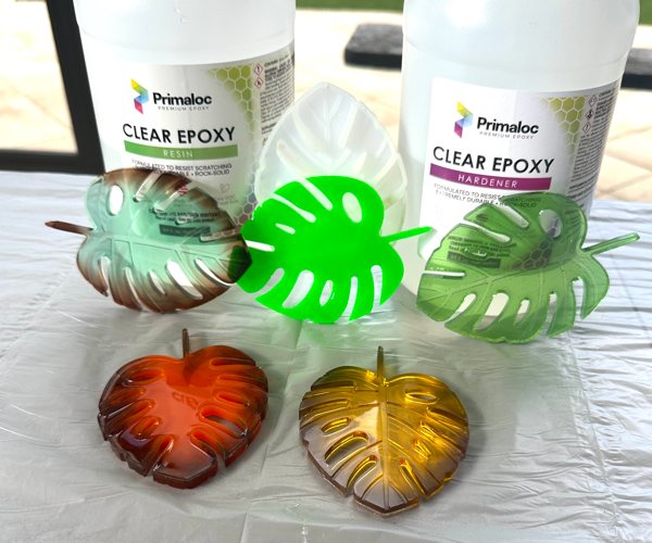 Five different leaf-shaped resin coasters resting in front of two bottles of Primaloc Epoxy. Each coaster is dyed with different colors.