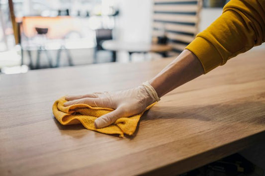 A gloved hand wiping down a wooden bar surface with an orange lint-free cloth.